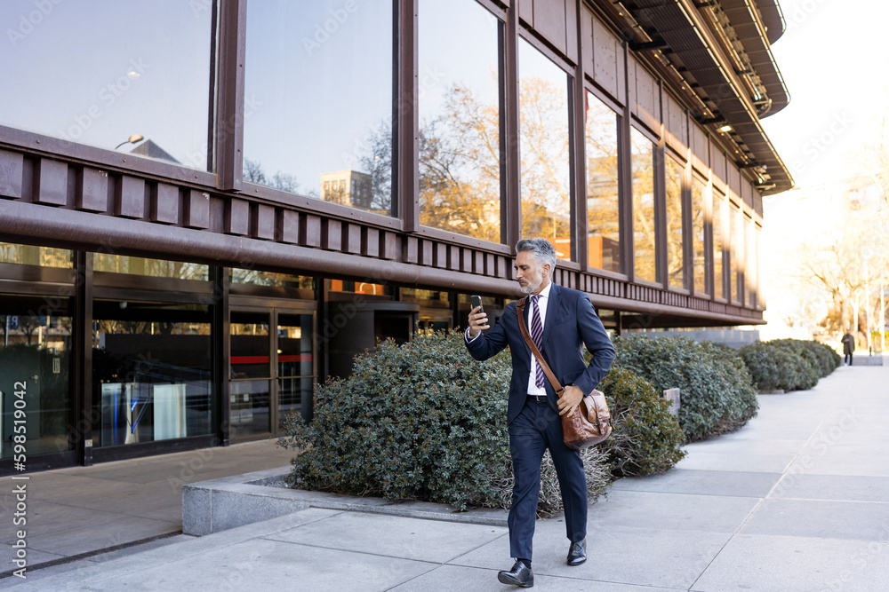 Businessman holding smart phone walking on footpath Businessman holding smart phone walking on footpath