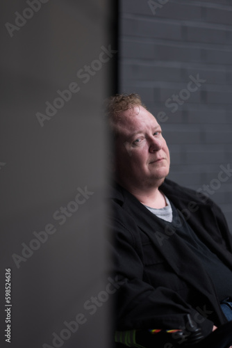 A man gives a slight smile, while posing in an entryway in a black wall. Adding backstory, he is trans and chronically ill, suggesting this photo also shows his pride and resilience. photo