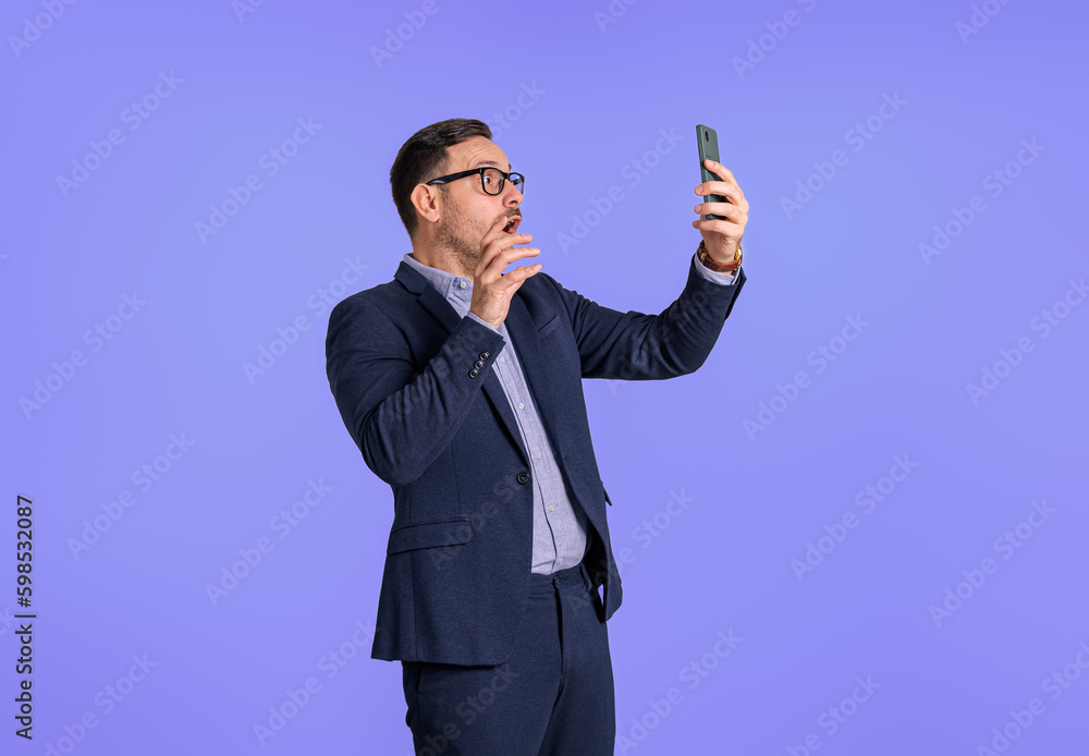 Displeased shocked young businessman dressed in suit reading messages on mobile phone. Worried male professional entrepreneur using cellphone while standing isolated on blue background Displeased shocked young businessman dressed in suit reading messages on mobile phone. Worried male professional entrepreneur using cellphone while standing isolated on blue background