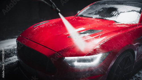 Car Wash Expert Using Water Pressure Washer to Rinse a Red Modern Sportscar. Adult Man Washing Away Dirt, Preparing an American Muscle Car for Detailing. Creative Cinematic Photo with Sport Vehicle photo