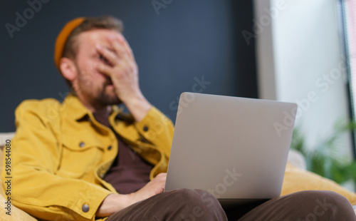 Man sitting on sofa with laptop, showing facepalm gesture with hand on face. Focus is on laptop, which suggests that the man may be experiencing frustration or disappointment related to the technology photo