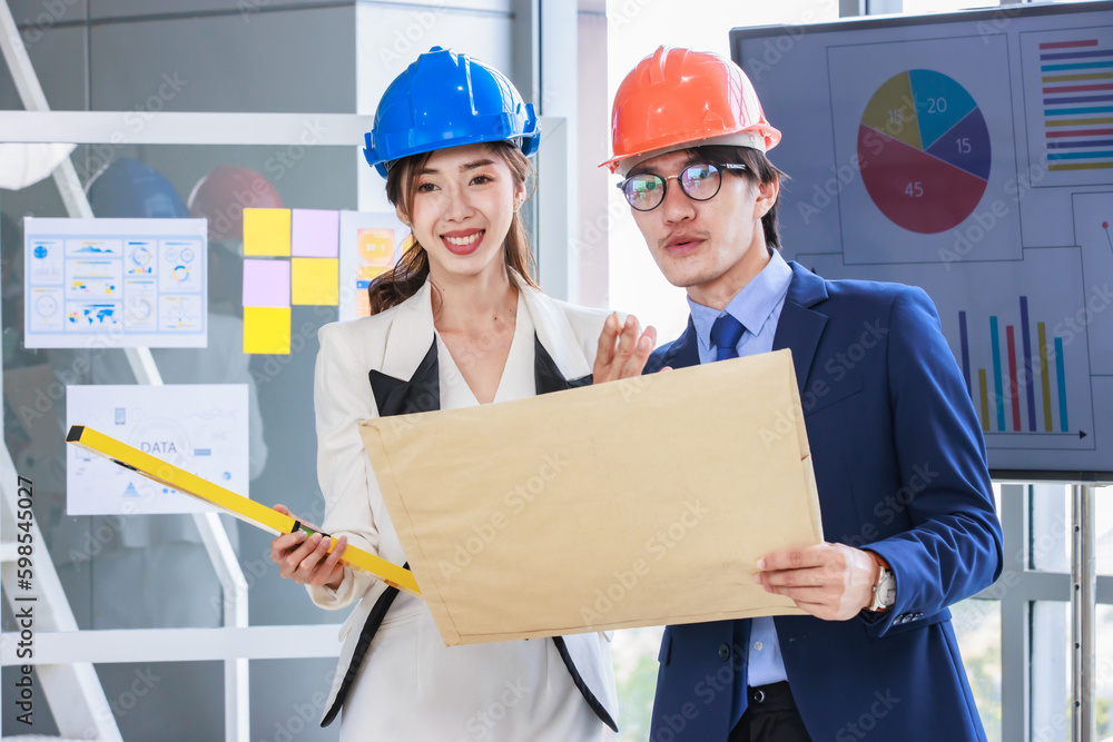 Asian professional successful female businesswoman and male businessman engineering employees in formal business suit and safety helmet standing holding blueprint and water level gauge discussing Asian professional successful female businesswoman and male businessman engineering employees in formal business suit and safety helmet standing holding blueprint and water level gauge discussing