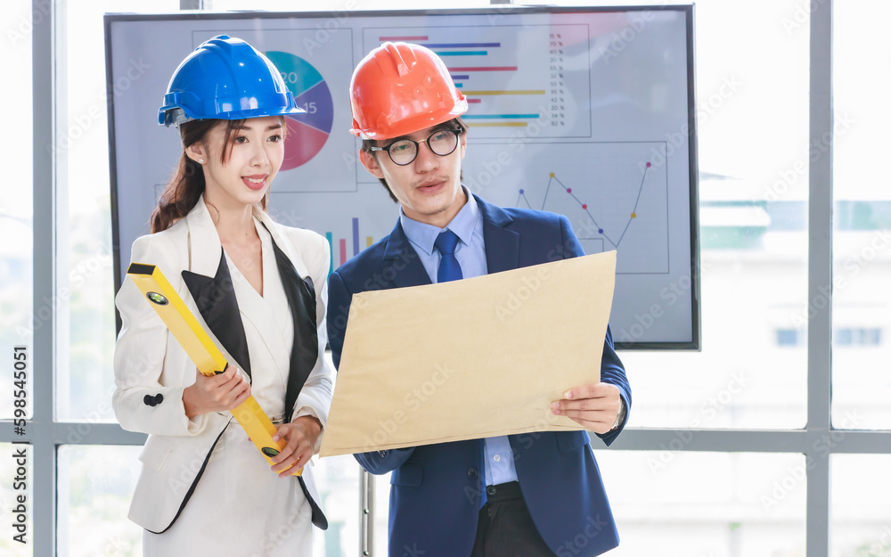 Asian professional successful female businesswoman and male businessman engineering employees in formal business suit and safety helmet standing holding blueprint and water level gauge discussing Asian professional successful female businesswoman and male businessman engineering employees in formal business suit and safety helmet standing holding blueprint and water level gauge discussing