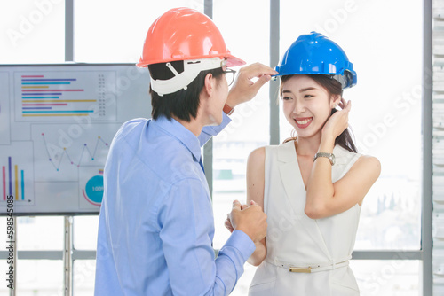 Asian professional successful male businessman holding water level gauge helping adjusting female businesswoman engineering safety hardhat standing smiling with document clipboard in meeting room