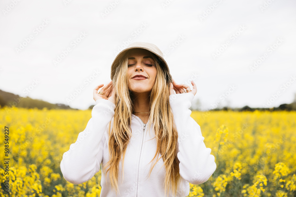 Beautiful young blonde cheerful woman, wearing a hat, poses amidst a field of blooming yellow rapeseed flowers Beautiful young blonde cheerful woman, wearing a hat, poses amidst a field of blooming yellow rapeseed flowers