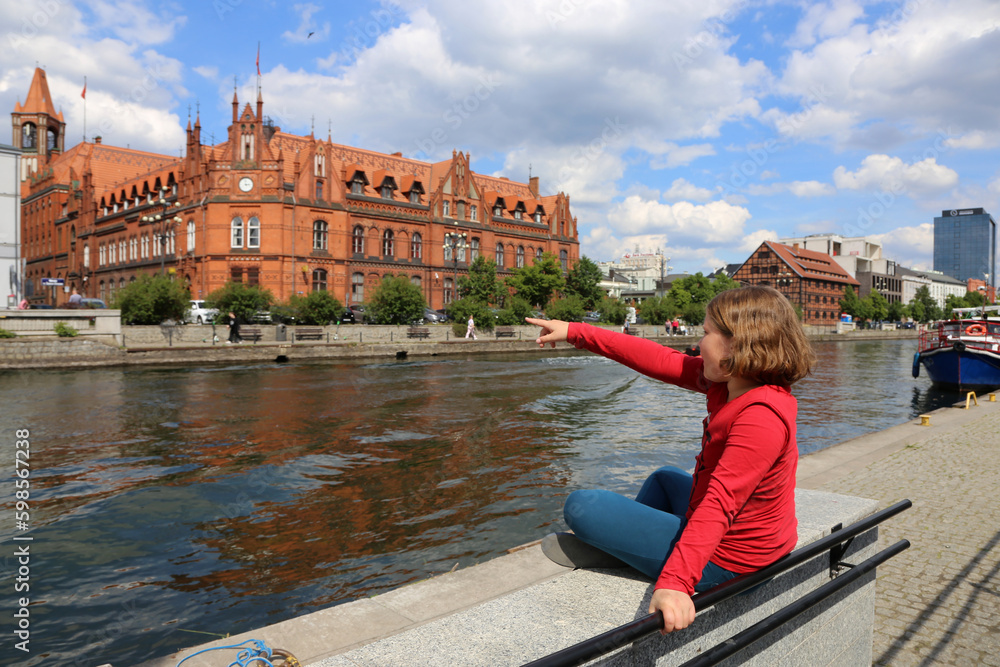 A girl sits on a parapet on the banks of the Brda River in Bydgoszcz. People visit riverfront in Bydgoszcz. Bydgoszcz is the capital of region Kujawsko-Pomorskie visited by 2.25m tourists annually  - obrazy, fototapety, plakaty 