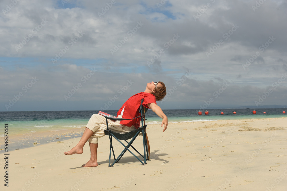 Mature woman relaxing on the beach and looking up to the sky. Senior woman wearing red shirt and sitting on the cheir. Active lifestyle after retirement. Side view