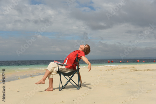 Mature woman relaxing on the beach and looking up to the sky. Senior woman wearing red shirt and sitting on the cheir. Active lifestyle after retirement. Side view © Nina