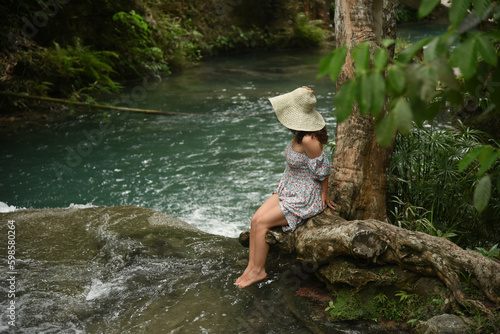Cute curly woman dressed in a dress and a straw hat sits near the river in the tropical jungle. Lonely girl tourist in the forest. Concept - travel around the world.