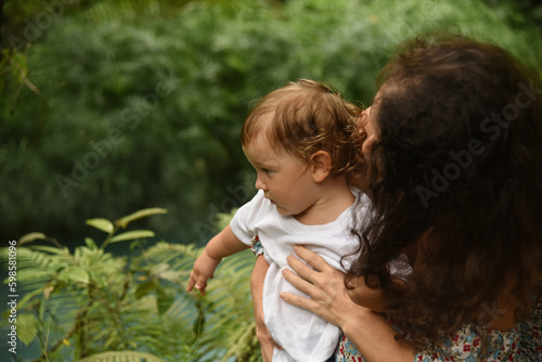 Thoughtful curly mother in a dress with a baby spend time in nature. Cute family in the summer city park. Mother hugs her son.
