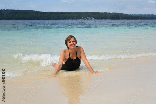 Happy attractive mature woman 50-60 lies in the water on the beach. Senior woman wear black swimsuit. Concept - travel  retirement  active elderly people  Philippines