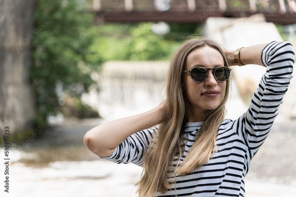 A woman in sunglasses and a striped top holds her hair near the river with her hands