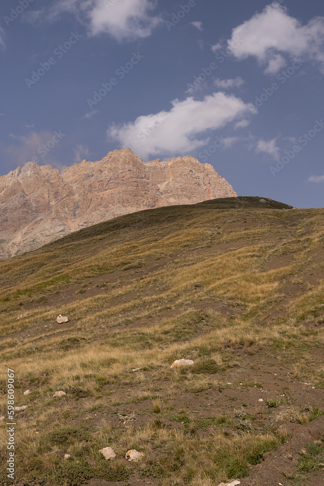 Mountain landscape - orange rocky bizarre cliff in bright sunny autumn day with clear blue sky and golden meadow with dry herbs on slope, vertical. Breathtaking tourism in Dagestan.