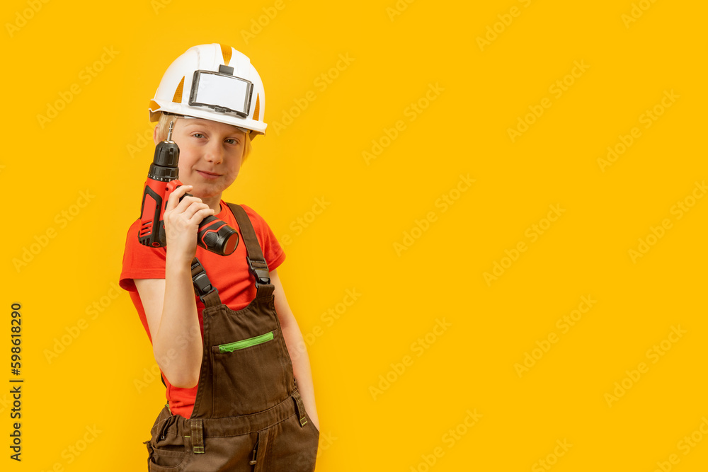 Child wears helmet and working overalls holds drill in his hands. Boy 9-10 years old imitates builder, yellow background. Copy space