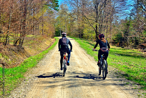 Back view photo of couple bikers  on forest road in spring sunny day. Unknown young friends exploring nature on bikes outdoors.  Training in spring forest. Weekend concept. Small depth of field. © Jurek Adamski