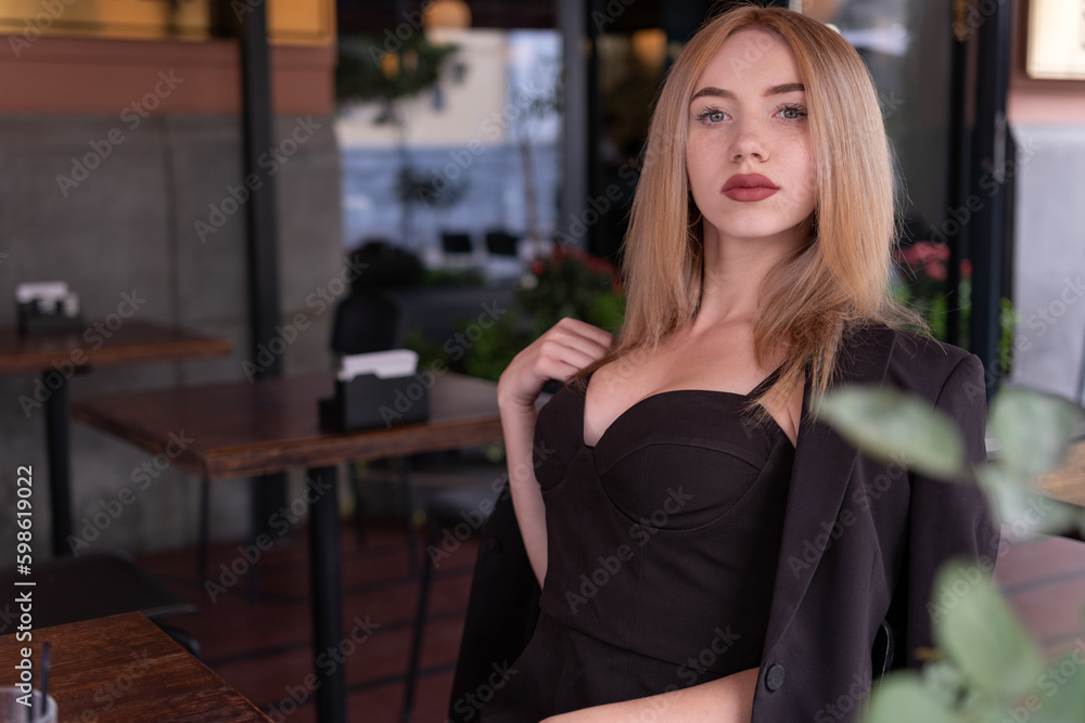 Sexy young woman with blonde hair and black suit with open neckline sits in cafe. Stares into the camera. Sexy young woman with blonde hair and black suit with open neckline sits in cafe. Stares into the camera.