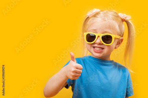 Cheerful preschool girl with blonde hair and two ponytails wears sunglasses. Child shows thumbs up, like gesture. Copy space.