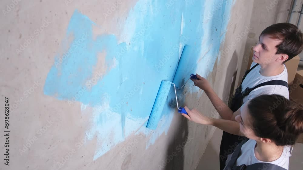 Couple in new home during repair works painting wall together. Happy family holding paint roller painting wall with blue color paint in new house. Home renovation DIY renew home concept Couple in new home during repair works painting wall together. Happy family holding paint roller painting wall with blue color paint in new house. Home renovation DIY renew home concept