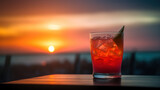 verre cocktail rouge avec une tranche de citron posé sur une terrasse qui domine la mer au couché du soleil
