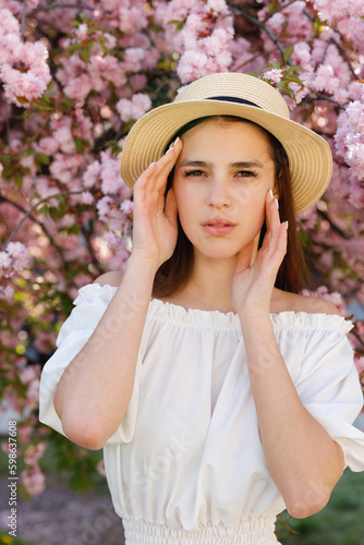Close Up. Cute Young Caucasian Dark Haired Girl in straw Hat Standing in the Park, Posing with Hands Beauty Poses, Near Sakura Tree With Pink Flowering. Femininity and Natural Beauty Concept