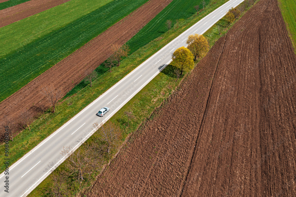 Obraz premium Aerial view of scenic countryside landscape in spring with country road and passenger car on it driving along on sunny spring day, drone pov.