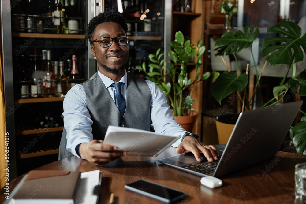 Portrait of young businessman in eyeglasses smiling at camera while working with laptop and document in cafe Portrait of young businessman in eyeglasses smiling at camera while working with laptop and document in cafe