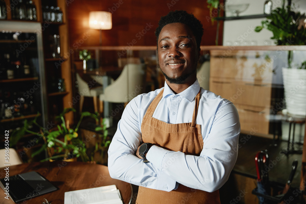Portrait of young African American waiter smiling at camera standing with his arms crossed and working in restaurant Portrait of young African American waiter smiling at camera standing with his arms crossed and working in restaurant