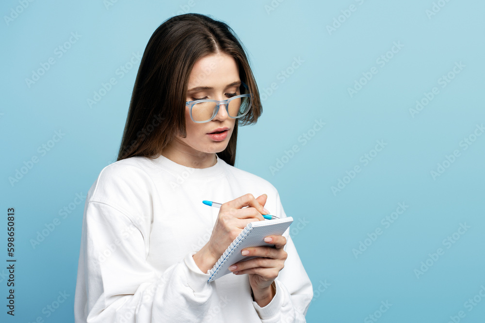 Smart student studying, learning language, exam preparation isolated on blue background, education concept. Pensive woman, writer wearing eyeglasses holding notebook, taking notes, copy space Smart student studying, learning language, exam preparation isolated on blue background, education concept. Pensive woman, writer wearing eyeglasses holding notebook, taking notes, copy space