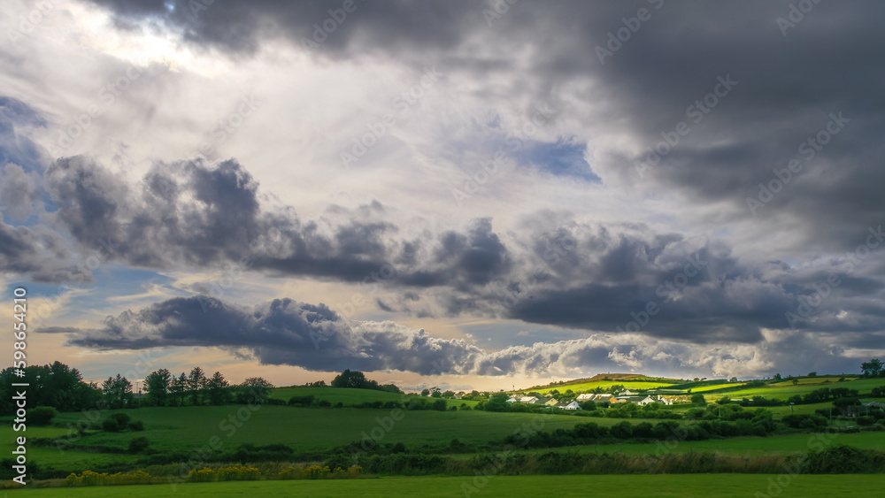 Sky with cumulus clouds over a Irish village on evening. Irish settlement in County Cork, dramatic landscape. European countryside, rustic landscape. Green grass field under cloudy sky