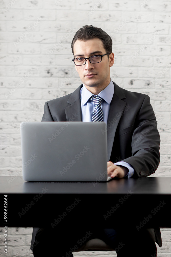 Handsome serious businessman in black suit sitting at the desk and working on laptop in the modern office Handsome serious businessman in black suit sitting at the desk and working on laptop in the modern office