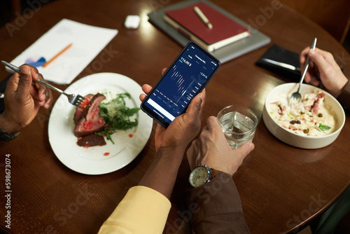 High angle view of business partners discussing financial graphs on the screen of smartphone during business lunch in cafe