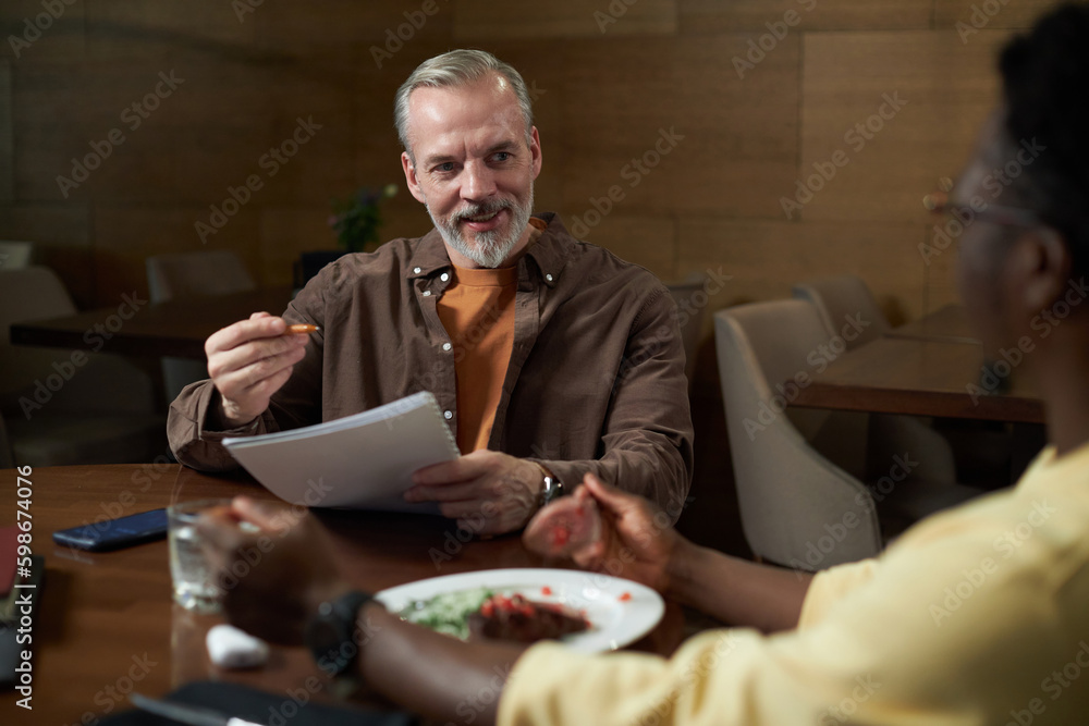 Mature businessman discussing terms of contract together with his colleague at meeting during business lunch in cafe Mature businessman discussing terms of contract together with his colleague at meeting during business lunch in cafe