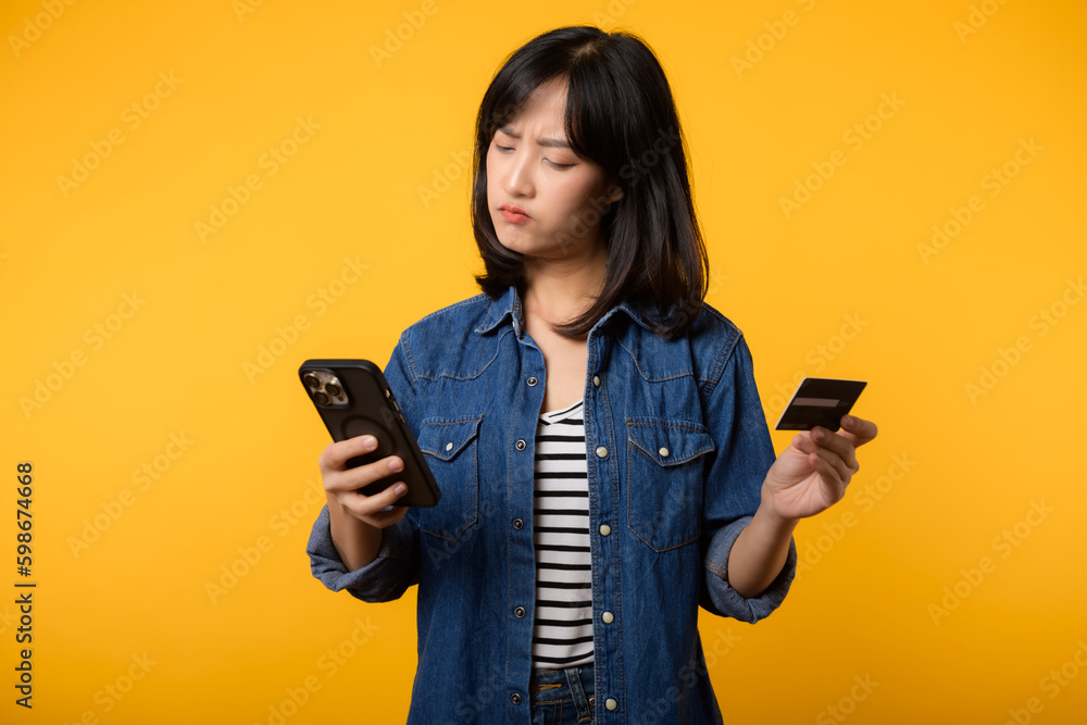 Portrait beautiful young asian woman happy smile dressed in denim jacket showing smartphone and credit card isolate on yellow studio background. Shopping online smartphone application concept Portrait beautiful young asian woman happy smile dressed in denim jacket showing smartphone and credit card isolate on yellow studio background. Shopping online smartphone application concept