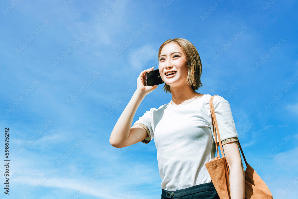青空をバックにスマホで話す笑顔の若い日本人女性 青空をバックにスマホで話す笑顔の若い日本人女性