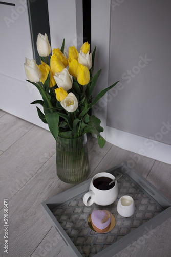 A vase with yellow and white near  a cup of coffee and cake. Modern room