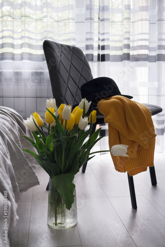 A vase with tulip flowers in a modern interior. A sweater and a hat on a chair in room - preparation for a date