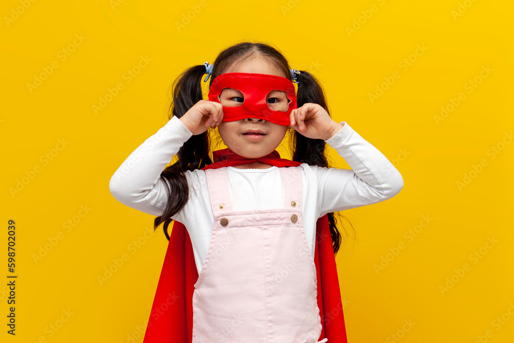 little asian girl in superman costume and mask, korean child in superhero cape smiling on yellow isolated background little asian girl in superman costume and mask, korean child in superhero cape smiling on yellow isolated background