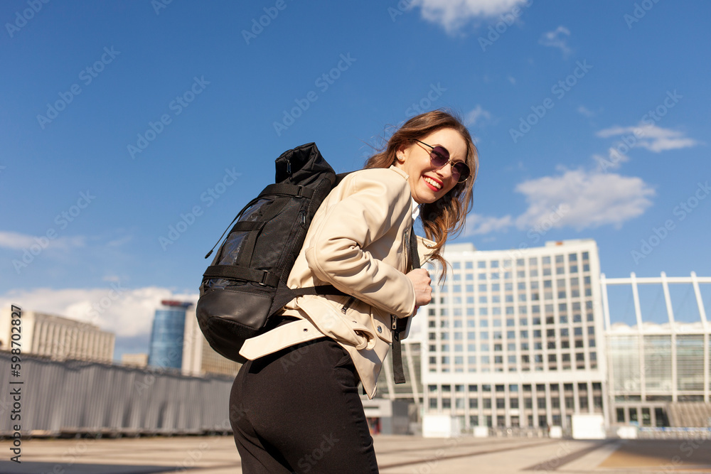 young happy girl with black backpack walks down the street in the city and smiles, woman in sunglasses travels