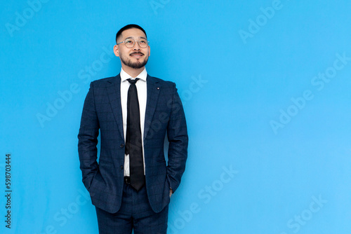successful asian businessman in suit stands on blue isolated background and smiles, korean entrepreneur in glasses