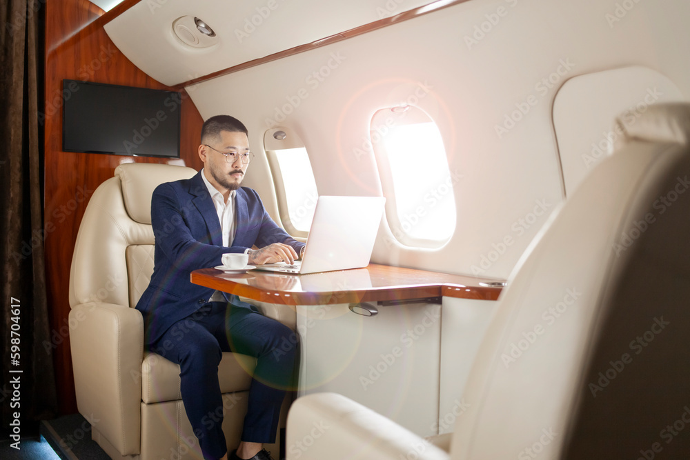 successful asian businessman in suit and glasses sits in private jet and types on laptop, korean entrepreneur