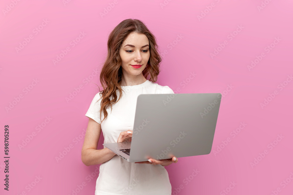 young cute woman in white t-shirt uses laptop on pink isolated background, woman freelancer types on computer young cute woman in white t-shirt uses laptop on pink isolated background, woman freelancer types on computer