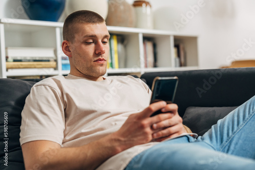 A handsome Caucasian man is sitting on the sofa and using his smartphone.