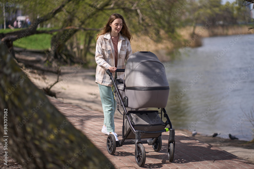 Young loving mother pushing gray baby stroller, slowly walking at town green park near a lake, in warm sunny spring day. Young loving mother pushing gray baby stroller, slowly walking at town green park near a lake, in warm sunny spring day.