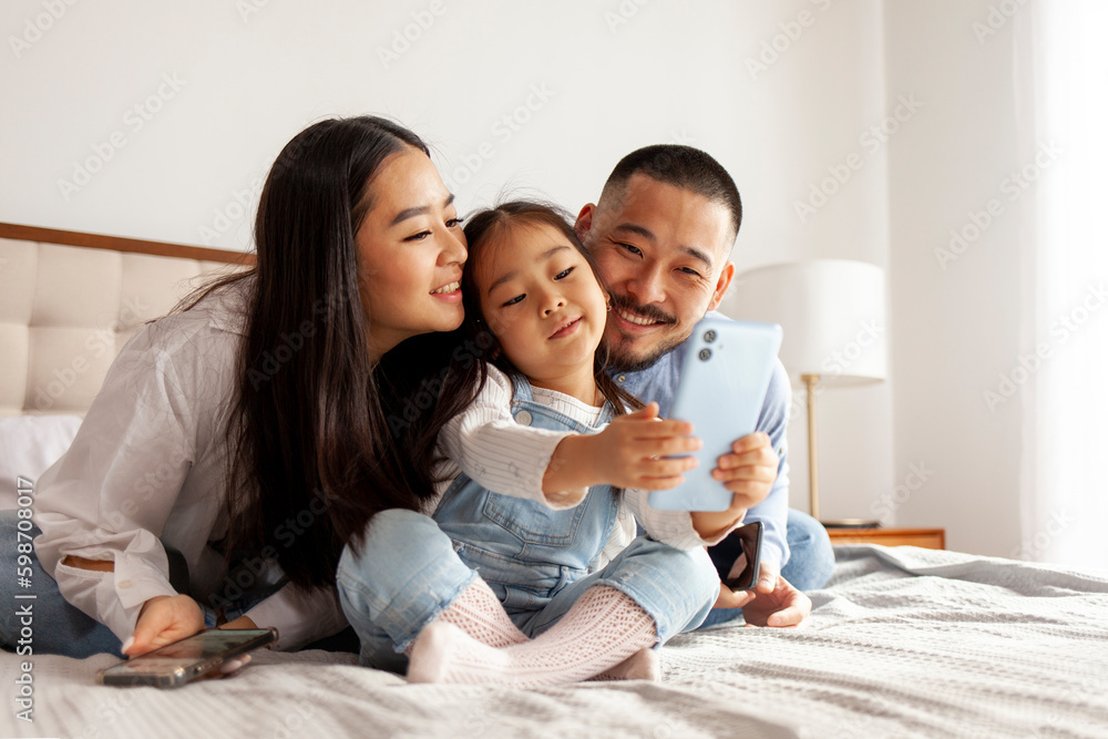 asian family sit on the bed and communicate via video call, little korean girl with mom and dad takes selfie asian family sit on the bed and communicate via video call, little korean girl with mom and dad takes selfie