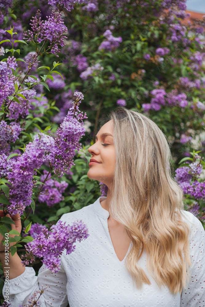 Fototapeta premium Portrait of beautiful young blue-eyed blonde woman near blooming lilac. Spring.