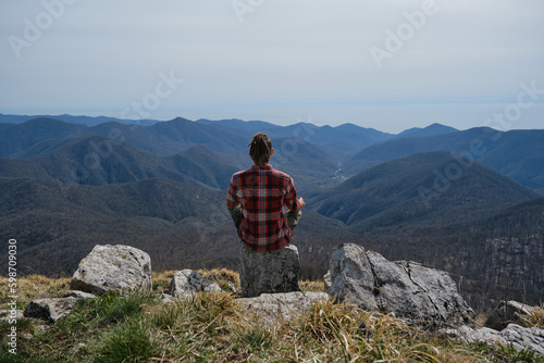 Guy traveler sitting on big rock in mountains and enjoying views of nature of Caucasus. Young man with dreadlocks meditates and does yoga outside. Rear view. Concept of travel and active lifestyle.