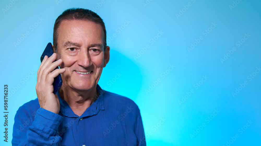 Happy and smiling mature adult caucasian male with blue shirt speaking on smartphone and looking at camera over pastel blue background with copy space Happy and smiling mature adult caucasian male with blue shirt speaking on smartphone and looking at camera over pastel blue background with copy space