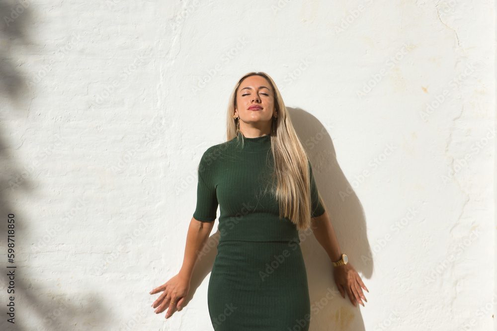 Young, beautiful, blonde woman with closed eyes, wearing a green dress, leaning against a white wall, relaxed, free and happy. Concept beauty, fashion, trend, eyes, happiness. Young, beautiful, blonde woman with closed eyes, wearing a green dress, leaning against a white wall, relaxed, free and happy. Concept beauty, fashion, trend, eyes, happiness.