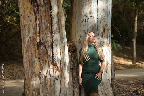 Young, beautiful, blonde woman with green eyes, wearing a green dress, leaning on the trunk of a big tree, with a dreamy look. Concept beauty, fashion, nature, autumn, dreams.
