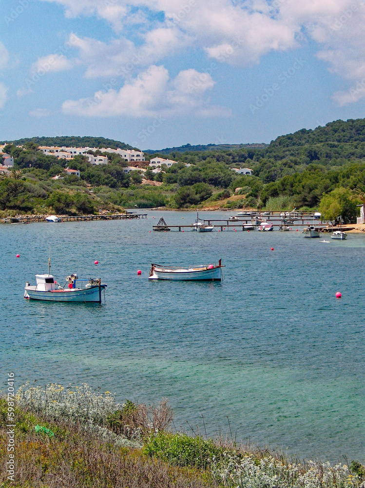 Obraz premium Scenic view over mediterranean sea with idyllic village and piers with mored boats at Spanish island Menorca on a sunny spring day. Photo taken May 18th, 2005, Menorca, Spain.